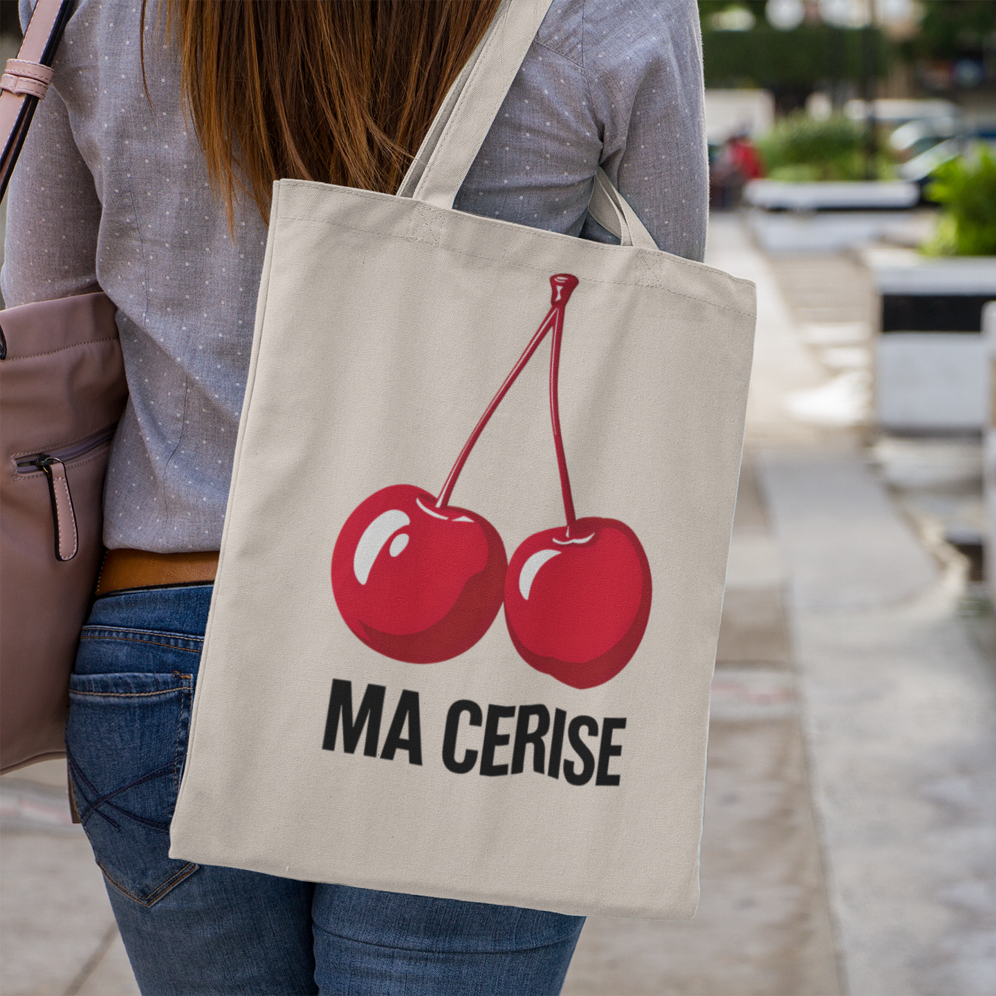 Person holding a tote bag with red cherries and 'MA CERISE' text in an outdoor setting.