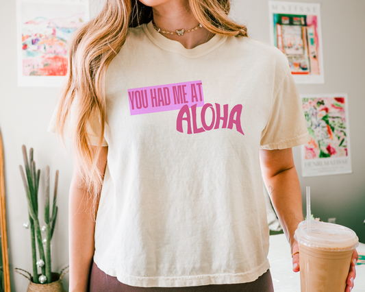Person wearing a white t-shirt with 'You Had Me at Aloha' text, holding a drink.