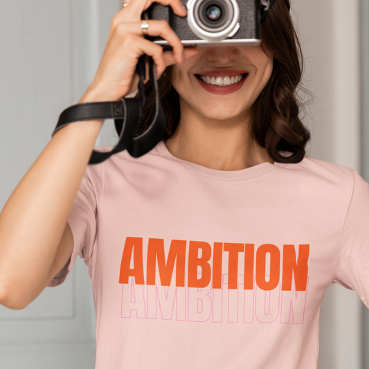 Person wearing a pink t-shirt with 'AMBITION' printed on it, holding a camera.