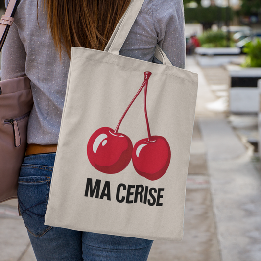 Person holding a tote bag with red cherries and 'MA CERISE' text in an outdoor setting.