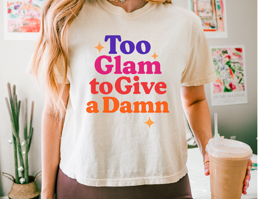 Person wearing a white t-shirt with colorful text 'Too Glam to Give a Damn' holding a drink.