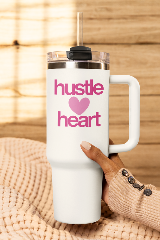 White tumbler with 'hustle heart' text and a pink heart, held by a person against a woven background.